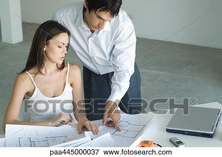 Stock Photo - Woman sitting at table looking at blueprints, man standing, looking over shoulder. Fotosearch
