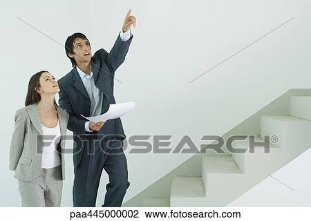 Young man and woman in suits standing at foot of stairs, man pointing upwards View Large Photo Image Stock Image - Young man and woman in suits standing at foot of stairs, man pointing upwards. Fotosearch