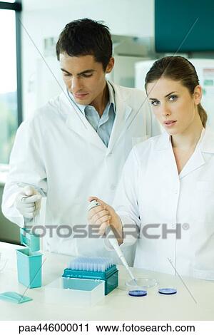 Stock Image - Male and female lab workers standing side by side, woman looking at camera. Fotosearch