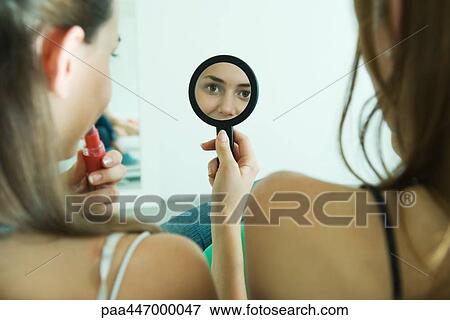 Stock Photo - Teenage girl sitting next to friend, applying lipstick, looking at self in hand mirror, rear view. Fotosearch