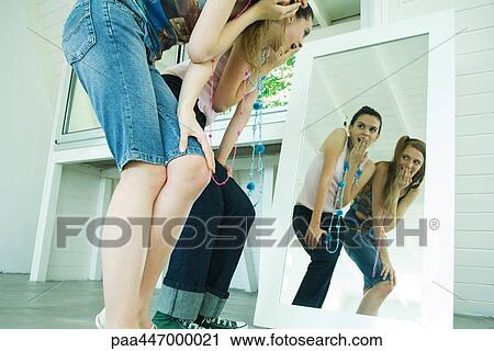 Stock Image - Two young friends bending over, looking at selves in mirror, covering mouths. Fotosearch