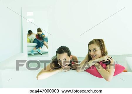 Stock Image - Two young friends leaning over back of couch, smiling at camera. Fotosearch