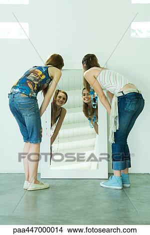 Picture - Two young friends looking at selves in mirror, bending over, rear view. Fotosearch