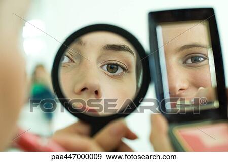 Stock Photo - Two young friends looking at selves in hand mirrors, applying lipstick, cropped view. Fotosearch