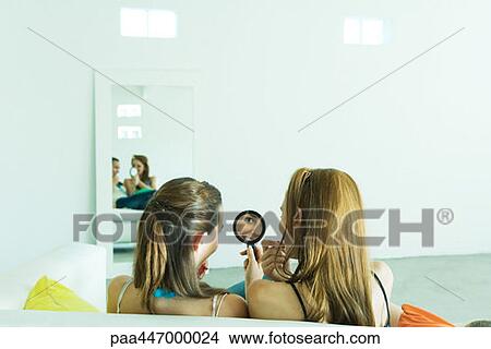 Picture - Two young friends on couch applying lipstick, looking in hand mirror, rear view. Fotosearch
