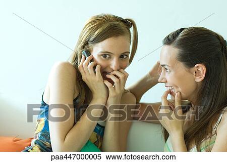 Stock Photography - Two young friends sitting together, smiling, one using cell phone, covering mouth. Fotosearch