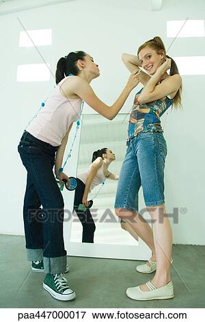 Stock Photo - Two young friends standing together, one leaning to apply lipstick to the other, one looking at camera. Fotosearch