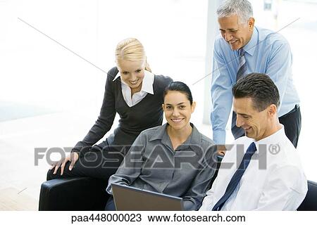 Stock Image - Business associates looking at laptop together, woman in center smiling at camera. Fotosearch