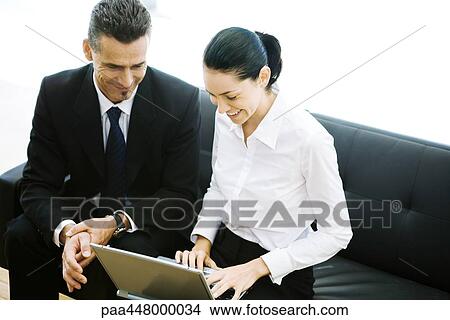 Picture - Male and female business associates sitting in lobby, using laptop, smiling. Fotosearch