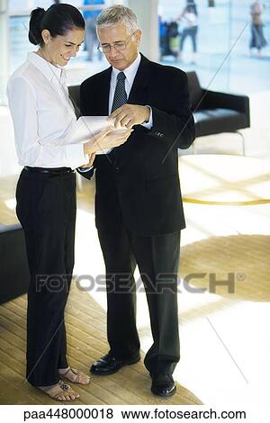 Mature businessman and young businesswoman standing, looking at file in office lobby, full length View Large Photo Image Stock Photo - Mature businessman and young businesswoman standing, looking at file in office lobby, full length. Fotosearch