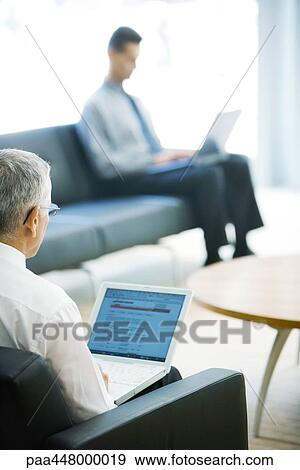 Stock Photo - Two businessmen using laptops in lobby, focus on mature man in foreground. Fotosearch