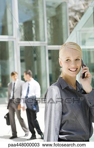 Stock Image - Young businesswoman using cell phone, looking away, associates in background. Fotosearch