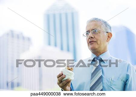 Stock Photo - Businessman holding sandwich, office buildings in background. Fotosearch