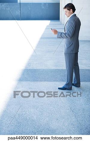 Businessman looking down at cell phone, full length View Large Photo Image Picture - Businessman looking down at cell phone, full length. Fotosearch