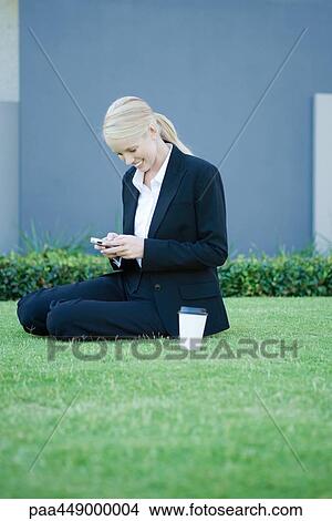 Businesswoman sitting on grass with coffee, looking at cell phone, smiling View Large Photo Image Picture - Businesswoman sitting on grass with coffee, looking at cell phone, smiling. Fotosearch