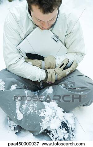 Stock Photo - Young man sitting in snow, embracing laptop computer, dressed in winter clothing. Fotosearch