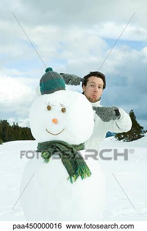 Stock Photograph - Young man standing behind snowman, pointing, making faces at camera. Fotosearch