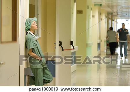 Stock Photo - Female nurse in hospital, leaning against wall with hands in pockets, side view. Fotosearch