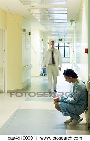 Stock Image - Male patient crouching in hospital corridor, looking down, doctor walking in background. Fotosearch