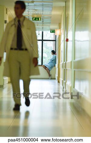 Picture - Man sitting, looking out window, wearing hospital gown, doctor walking in foreground. Fotosearch