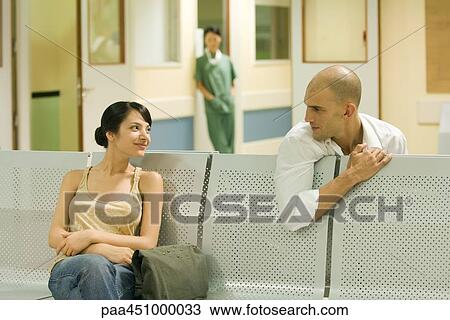 Stock Image - Two adults sitting in hospital waiting room, chatting. Fotosearch