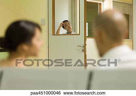 Stock Photograph - Woman looking at camera through window in hospital waiting room, adults in foreground. Fotosearch