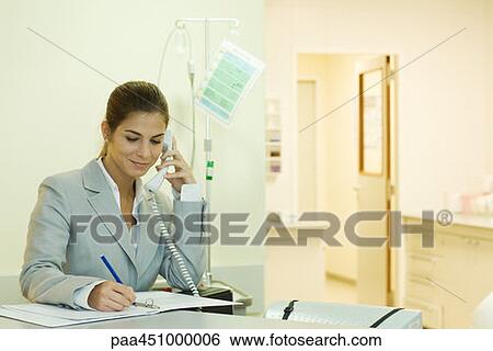 Stock Photograph - Young woman in doctor's office, using phone, writing. Fotosearch