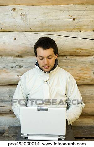 Picture - Young man using laptop next to log cabin. Fotosearch