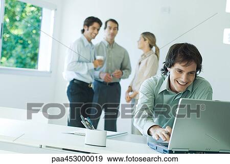 Businessman in office, using laptop computer, smiling, associates in background View Large Photo Image Stock Photo - Businessman in office, using laptop computer, smiling, associates in background. Fotosearch