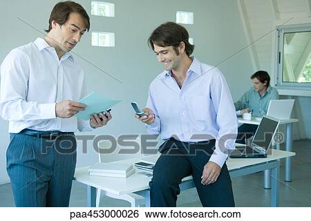 Businessmen in office, one using cell phone, smiling View Large Photo Image Stock Photograph - Businessmen in office, one using cell phone, smiling. Fotosearch