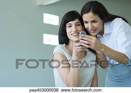 Two businesswomen looking at cell phone, smiling, cheek to cheek, waist up View Large Photo Image Stock Photo - Two businesswomen looking at cell phone, smiling, cheek to cheek, waist up. Fotosearch