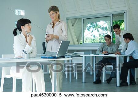 Two young businesswoman in office, chatting, businessmen in background View Large Photo Image Stock Photography - Two young businesswoman in office, chatting, businessmen in background. Fotosearch