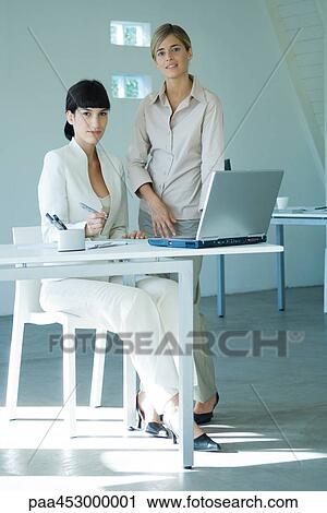 Stock Image - Two young businesswomen in office, smiling at camera, full length. Fotosearch
