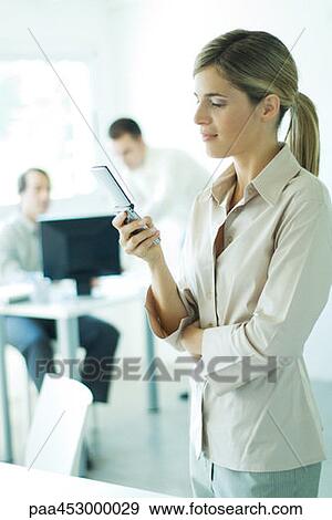 Stock Photo - Young businesswoman in office, using cell phone, associates in background. Fotosearch