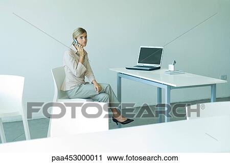 Stock Image - Young businesswoman seated in office, using cell phone, full length. Fotosearch