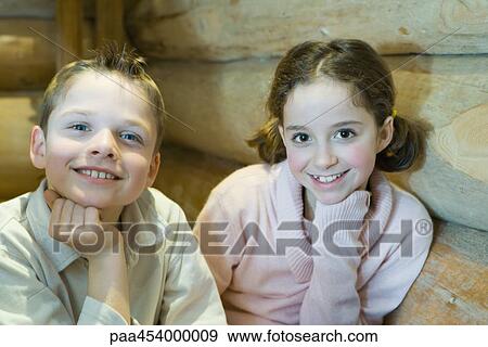 Stock Photo - Boy and girl sitting side by side, hand under chin, smiling at camera. Fotosearch