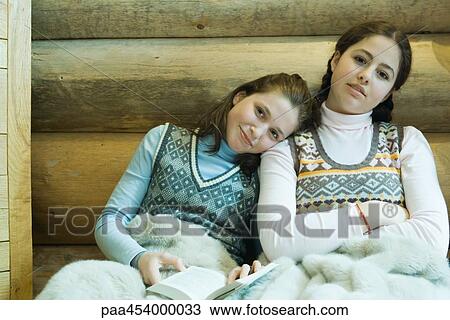 Stock Image - Two teenage girls sharing blanket, smiling at camera, one holding book, resting head on the other's shoulder. Fotosearch