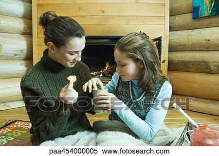 Stock Photography - Two teenage girls sitting by fireplace, sharing snack. Fotosearch