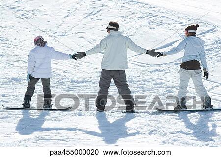 Stock Image - Three young snowboarders standing together, holding hands, rear view. Fotosearch