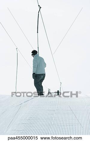 Stock Photography - Young skier riding ski lift, rear view. Fotosearch
