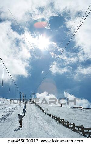 Stock Image - Young skiers using ski lift on hill. rear view, in the distance. Fotosearch