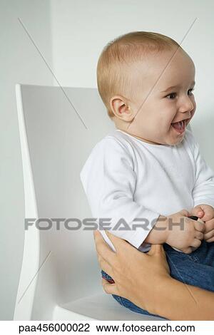 Stock Image - Baby sitting, looking away, cropped view of woman's hands. Fotosearch