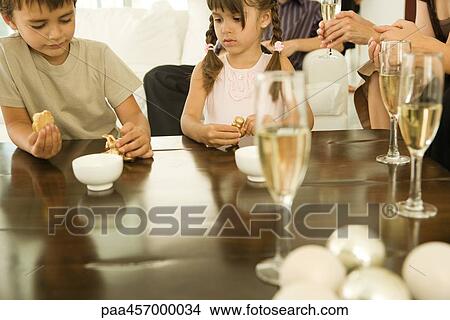 Boy and girl sitting at table, both holding Christmas ornaments, adults drinking champagne in background View Large Photo Image Picture - Boy and girl sitting at table, both holding Christmas ornaments, adults drinking champagne in background. Fotosearch