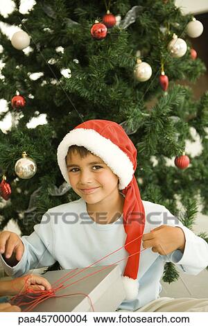 Picture - Boy opening Christmas present in front of Christmas tree, wearing Santa hat, smiling at camera. Fotosearch