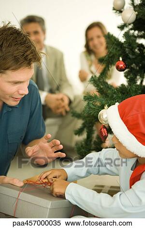 Stock Photography - Father and son opening Christmas present in front of Christmas tree, boy wearing Santa hat. Fotosearch