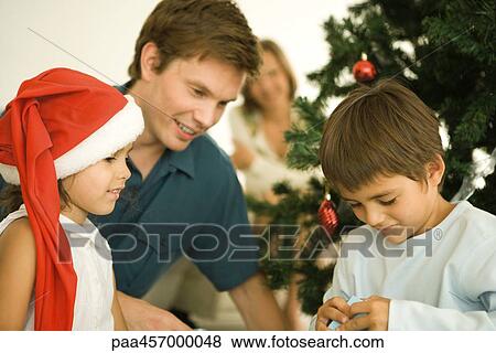 Father and two children opening Christmas present in front of Christmas tree, girl wearing Santa hat View Large Photo Image Stock Photo - Father and two children opening Christmas present in front of Christmas tree, girl wearing Santa hat. Fotosearch
