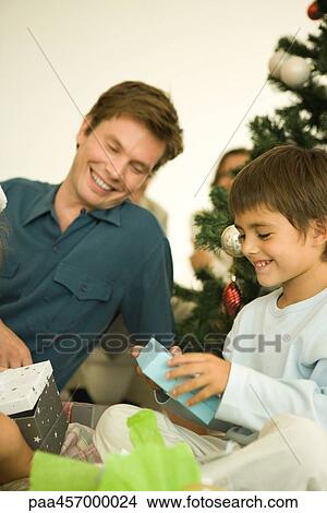 Man and boy sitting by Christmas tree, opening Christmas present, smiling View Large Photo Image Picture - Man and boy sitting by Christmas tree, opening Christmas present, smiling. Fotosearch