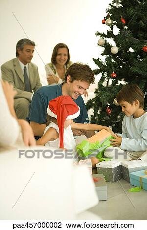 Stock Image - Man and two children opening Christmas presents in front of Christmas tree, family watching in background. Fotosearch