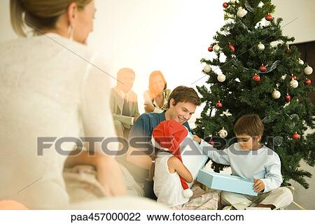 Stock Image - Man and two children opening Christmas present in front of Christmas tree, woman watching in foreground. Fotosearch