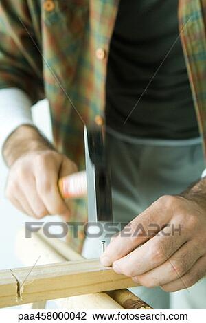 Stock Image - Man hammering nail into wood, cropped view. Fotosearch